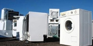 A group of white washing machines and refrigerators are stacked on top of each other, awaiting junk removal.