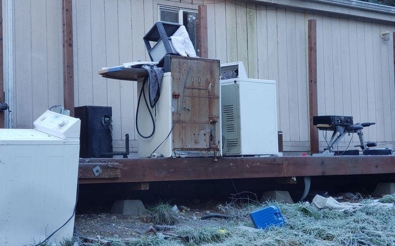 A refrigerator and microwave, along with various other appliances, are resting on a wooden deck. The need for trash removal WA services might arise to dispose of any unwanted items.